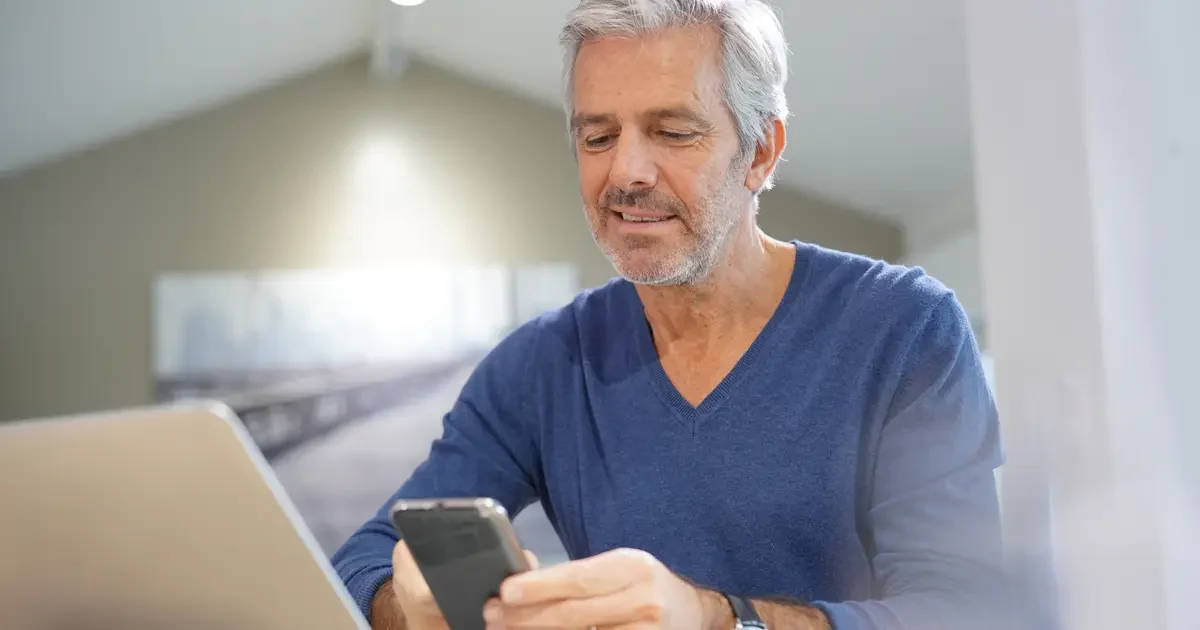 Smiling man uses smartphone at table with laptop in modern home.