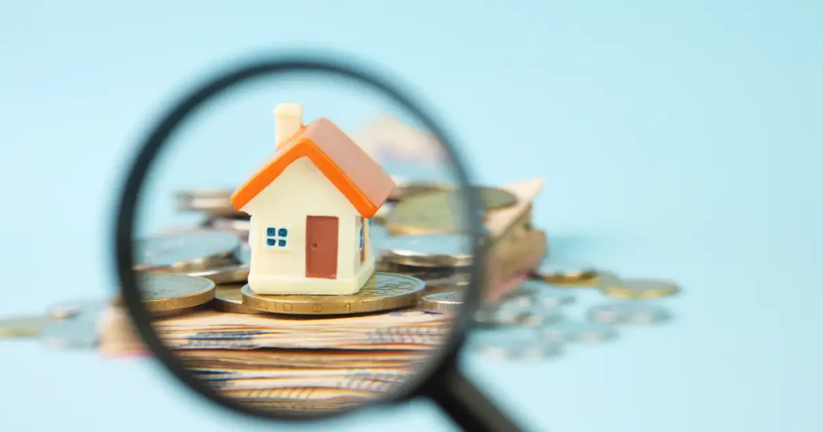 Miniature house on money stack under magnifying glass.