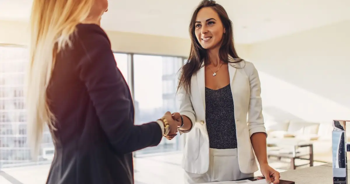 Two professional women shake hands in modern office, smiling confidently.