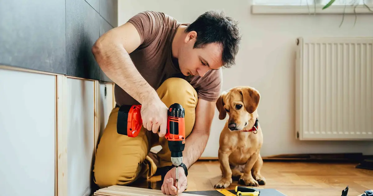 Man using power drill during renovation as dog watches beside him.