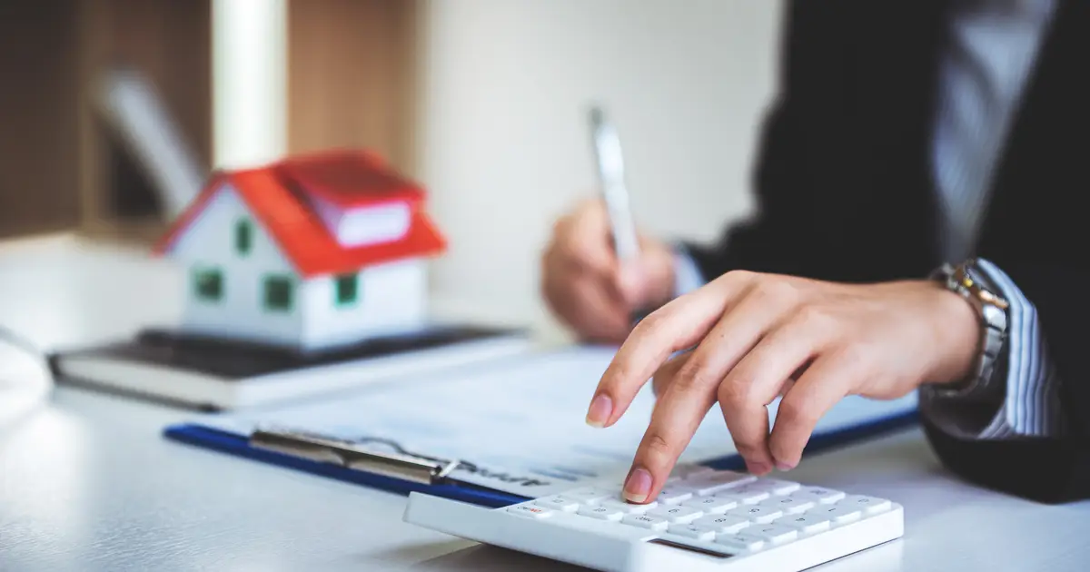Person calculates mortgage with documents and model house on desk.